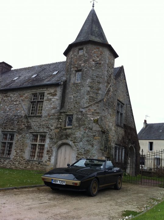 wedge trip to france - Page 1 - France - PistonHeads - The image prominently features a large, old, multi-story building constructed of stone. Its architecture is characteristic of a historic or medieval style, with a castle-like structure and a sturdy gate. Parked in front of the building is a classic, black convertible car. The sky is overcast, and the ground appears to be made of gravel. In the background, there's another structure partially visible, suggesting that the main building might be part of a larger complex or property. Overall, the image captures a scene that evokes a sense of history and elegance due to the combination of the classic car and the historic building.