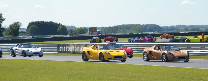 Snetterton Lotus Festival 25/26 June - Page 1 - General Lotus Stuff - PistonHeads - The image captures a thrilling scene on a race track. A group of sports cars, in vibrant hues of yellow, red, and white, are spearing ahead in a race. They are driving on the left side of the road, their configurations mirroring those on the wider track they occupy. The track is surrounded by lush green scrubs and verdant trees under a clear blue sky. The close-up perspective of the image suggests it was taken from the sidelines, adding a sense of excited anticipation to the scene.