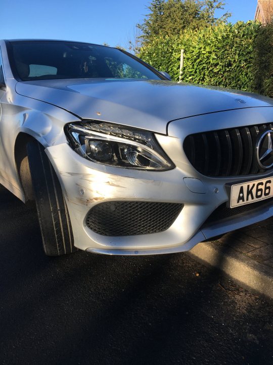 Idiot crashed into me - Page 1 - General Gassing - PistonHeads UK - The image shows a white Mercedes-Benz vehicle parked on the side of a road. The car appears to have been in an accident, as evidenced by the visible damage and scratches on its front bumper and hood. There's a noticeable dent in the passenger door area. A small part of a person is partially visible at the edge of the frame, possibly suggesting the presence of someone inspecting or examining the car. The vehicle has a European license plate with UK style registration letters and numbers.