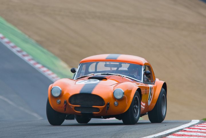 Pistonheads - In the image, a vibrant orange classic race car is the focal point. The vehicle is predominantly orange with black accents, giving it a distinctive racetrack aesthetic. The car is moving at high speed on a curved track, indicating the dynamic nature of the scene. The driver's hands can be seen gripping the steering wheel as they navigate the tight curve, adding a sense of action and excitement to the image. The background appears to be a specially designed race track with a dirt surface, emphasizing the performance capabilities of the car.