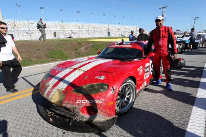 Daytona 24hr tour  - Page 1 - Vipers - PistonHeads - In the image, a scene on a race track is depicted. A person dressed in red clothing is standing next to a red and white car. The car appears to have some wear and tear but is dominated by these two colors. The person is standing in the foreground, positioned parallel to the car as if posing for a photo. The race track setting is confirmed by a blurred race car in the background, implying motion, a common sight at such venues. Overall, the image captures a moment of pause in the excitement of a racing event.