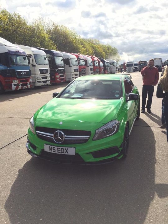 The not so common A45 AMG - Page 3 - Readers' Cars - PistonHeads - The image features a vibrant green Mercedes-Benz MS5 E-class car parked on a concrete surface. The vehicle bears a UK registration plate and is facing towards the left. The car's striking color stands out against the overcast sky. In the distance, parked behind the car, are several large commercial trucks, predominantly red and white. The scene takes place during the day under a uniform cover of clouds. People can be seen standing near the trucks, perhaps engaged in conversation or preparing for a journey.