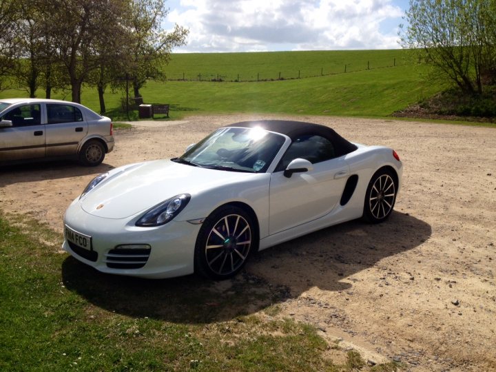 20 inch wheels without PASM - Page 1 - Boxster/Cayman - PistonHeads - The image depicts a scene of a white Porsche parked on a gravel driveway. The car is positioned in profile, showcasing its design. The sky above is a bright blue, indicating fair weather. To the side of the driveway, there's a tree, and in the background, there's a carport starkly contrasting with the bright surroundings. The overall scene suggests a serene and peaceful setting.