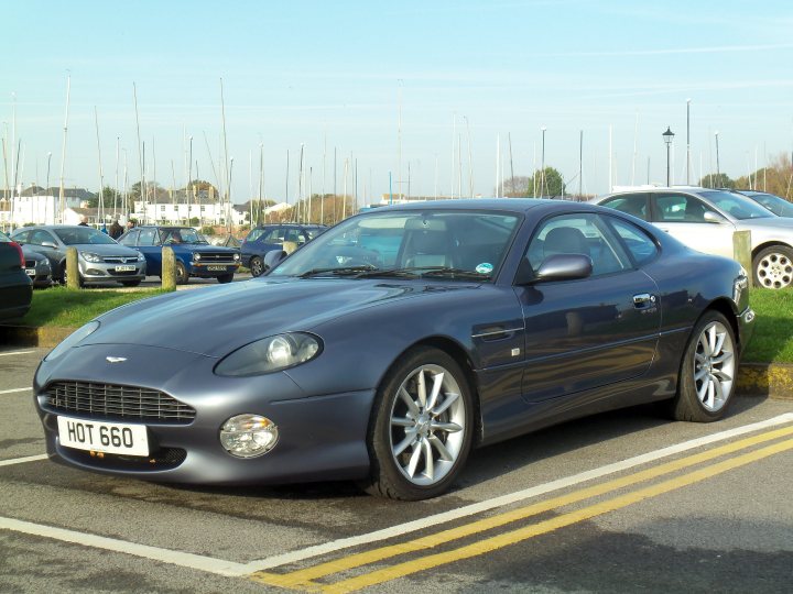 Show us your DB7 .... - Page 5 - Aston Martin - PistonHeads - The image depicts a blue Aston Martin sports car parked in a parking lot with various other cars and a few pedestrians. The Aston Martin is parked alongside a yellow dividing line. In the background, there is a building, a few parking signs, and a grassy area. The sky suggests it's a clear day, and the overall scene is calm and everyday, with the Aston Martin being the focal point due to its bold color and luxury status.