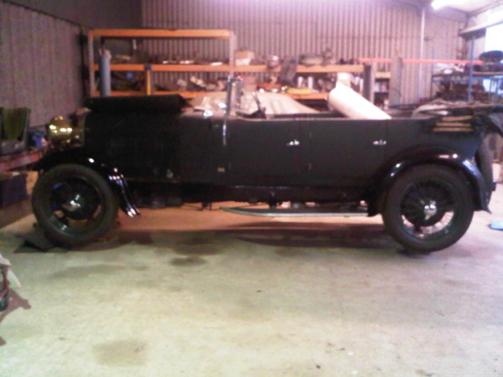 Lagonda Thread - Page 1 - Aston Martin - PistonHeads - The image shows a black vintage motor car parked in a garage. The car appears antique, with a streamlined body, chrome details, and shiny black tires. The garage is filled with age-old tools and parts on shelves and on the floor, suggesting that it might be a space for restoration or repair work on vintage vehicles. The atmosphere is one of nostalgia and mechanical history.