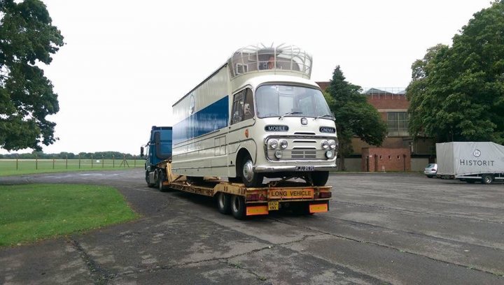 The Heavy Haulage Appreciation Thread - Page 12 - Commercial Break - PistonHeads - This image depicts an oversized truck in transit on the left side of the road. The truck, taking up both lanes, appears to be a two-story vehicle with multiple windows lining its upper body, possibly designed for long-haul travel. Its bed is visible, holding up the back of the two-story structure. The truck is backed by a blue trailer and is pulling a movable flatbed trailer with a van placed on top. The flatbed trailer has a license plate that reads "LONG WHILE". The flatbed trailer is also bearing another license plate, which is partially visible. The background features a gray sky and trees, indicating a suburban or rural area.