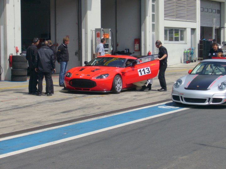 New Zagato - Page 1 - Aston Martin - PistonHeads - This image captures an exciting scene on a track. There's a race between two sports cars, one red and the other gray. Multiple people can be seen inspecting the red car, which is numbered 113. Behind the vehicles, there's a white building typical of racing or automotive facilities. The atmosphere is of anticipation and adrenaline, characteristic of car racing.