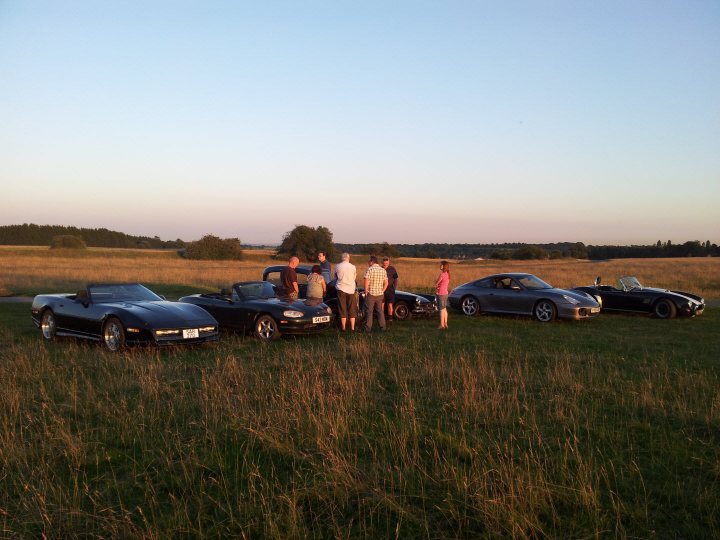 A group of people standing around a truck - Pistonheads - The image depicts a tranquil setting of a grassy field under the soft glow of a sunset. There are three vintage cars parked haphazardly on the tall, dry grass. A group of people, approximately six, are standing around these cars. They appear to be observing the cars from different angles. The sky is speckled with soft hues as the day transitions to night. The overall vibe of the scene is one of camaraderie and enjoyment of the classic cars among the friends.