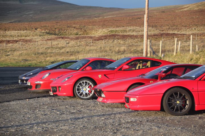 Great drive out today to Hartside Cafe - IN NOVEMBER !!! - Page 1 - Supercar General - PistonHeads - This image features a collection of red sports cars parked on a gravel lot. The cars are all similar in make, likely Ferrari, and they are parked side by side. One car is seen with the door open, suggesting that someone might be inside. The backdrop of the scene is a vast grassy field reaching into the distance, and in the far distance, patches of trees can be seen. The overall setting conveys a sense of tranquility, with the colorful, high-performance vehicles contrasting against the natural landscape.