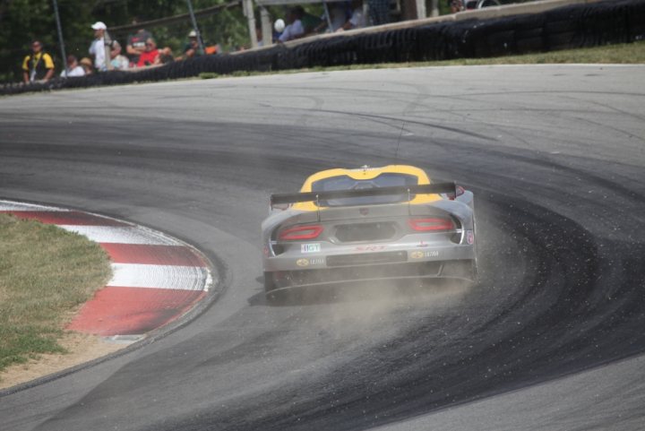 Pistonheads - The image captures a dynamic scene from a racing track. A sports car, numbered 69 and sponsored by VMI, is driving at high speed, leaning into a sharp left bend known as a chicane. The vehicle sports a predominantly yellow body with a gray roof, and black and red details. The setting sun casts long shadows, enhancing the sense of speed and motion. Fans can be seen watching the race from the edge of the track, adding to the atmosphere of the event.