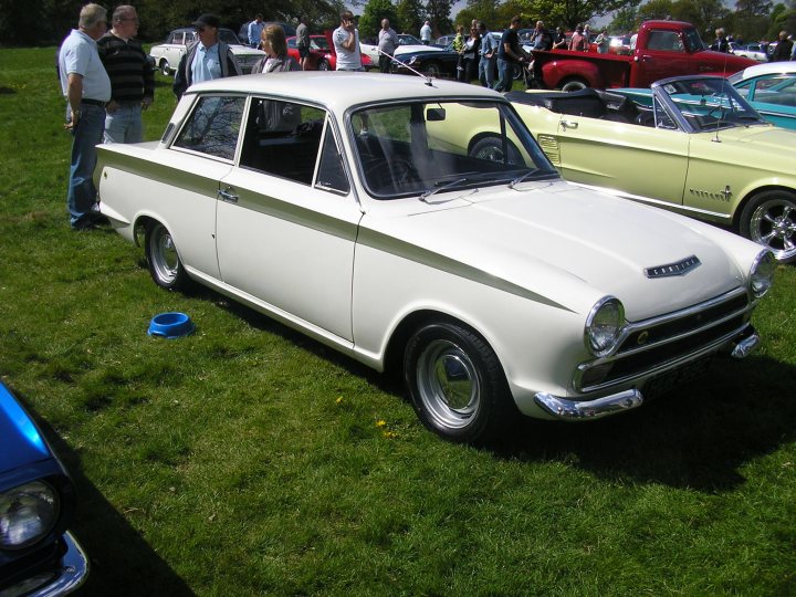 June Classic Pistonheads Ford - The image captures a classic white car parked in the grass at a car show. The car features a distinctive white and black hood ornament and stands out against the greenery. In the background, various other cars, including a vivid red one, are also displayed on the grass, adding to the atmosphere of a classic car gathering. The attendees, a small group of people, cluster around the cars, engaged in conversation, enjoying the display of vintage automobiles.