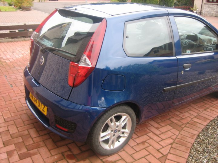 My first car! - Page 1 - Readers' Cars - PistonHeads - The image shows a small dark blue car parked on a driveway made of light-colored bricks. The vehicle has a hatchback body style, characterized by a prominent rear hatch. The car features a spare tire mounted on the back of the vehicle, and there is a license plate visible on the back bumper. The image itself is a bit blurry, indicating either the camera was moving or the car was slightly out of focus.