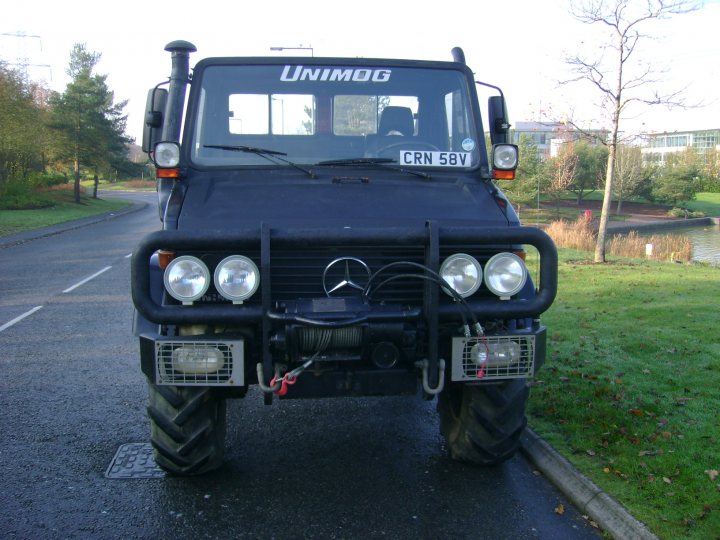 A truck that is sitting in the street - Pistonheads - The image displays a dark green all-terrain vehicle (ATV) sitting on a paved road. The vehicle is branded with "UNIMOG" and the registration number "CRN 59V". It has a front-mounted spare wheel, and there are two white lights visible on the front bumper. In the background, there are trees and what appears to be a body of water, possibly a lake or pond. The vehicle is parked and there are no other vehicles immediately visible.