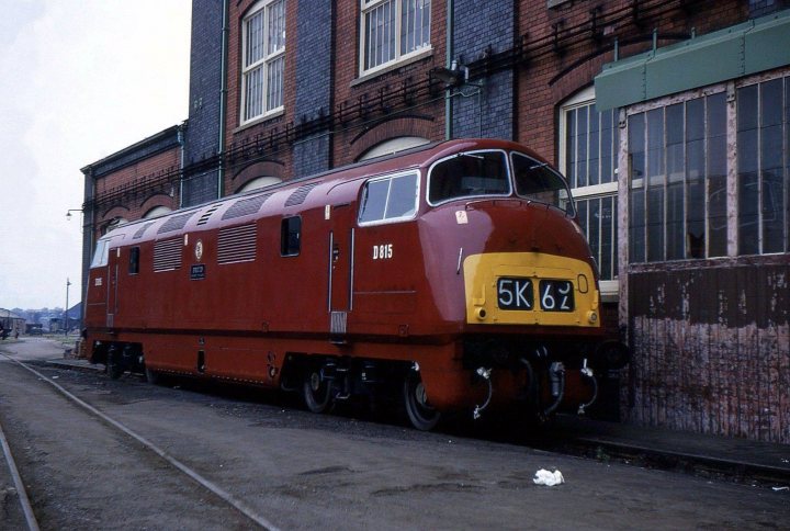 Loco sheds and other railway buildings.... - Page 16 - Boats, Planes & Trains - PistonHeads UK - This image features a vibrant red diesel locomotive parked on tracks next to a building. The locomotive has the number "867" prominently displayed. There is a solitary person standing near the locomotive, possibly an engineer or a railway worker. In the background, a brick wall and a metal structure are visible, suggesting an industrial setting. The overall scene conveys a sense of anticipation for a journey about to begin.