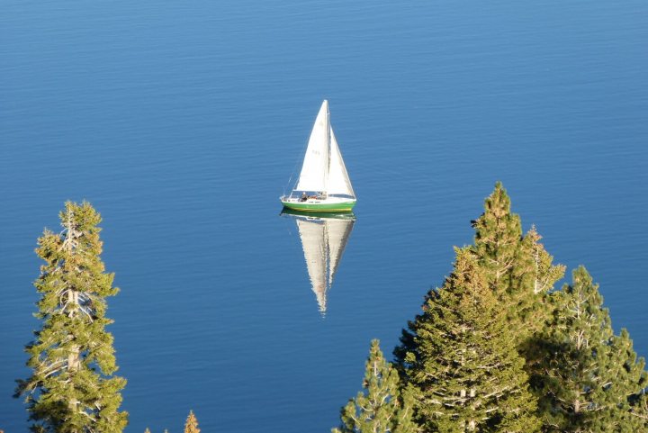 A boat floating on top of a body of water - Pistonheads - In the tranquil setting of a blue-green lake, a green sailboat is gracefully sailing towards the right side of the image. The boat, with its white sail spread wide, is the main focus of this serene landscape. The lake is encircled by tall evergreen trees, lending a forested ambiance to the scene. The boats reflection dances on the water's surface, creating a peaceful and inviting atmosphere. The image beautifully captures a moment of simple joy and serene solitude.