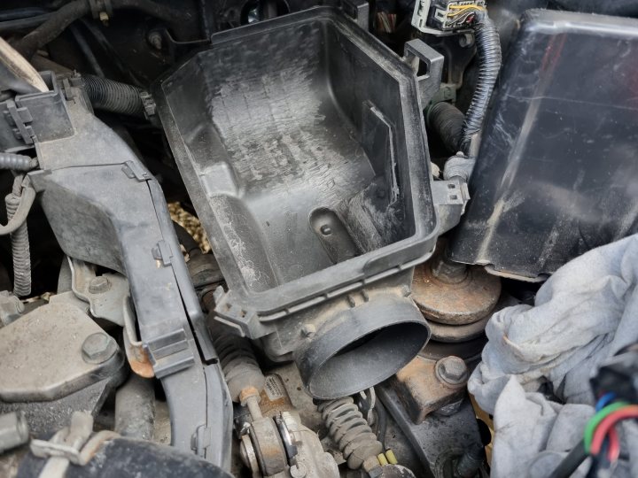 Civic Type R FN2 Track Car  - Page 3 - Readers' Cars - PistonHeads UK - The image shows a vehicle's engine compartment with various components exposed. On the left, there appears to be an electrical connection, and some wiring can be seen. In the center of the image, there's a large air intake cover with visible screws. To the right, several parts are scattered around, including what seems to be an engine cover lying on its side and a black plastic casing. The engine block itself is not fully visible, but it's clear that the vehicle has undergone some disassembly or repair work. The surrounding area looks like a garage or workshop environment, as suggested by the presence of a toolbox and other mechanical equipment in the background.