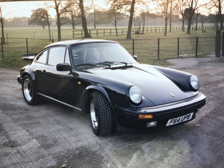 Pictures of your classic Porsches, past, present and future - Page 46 - Porsche Classics - PistonHeads - The image shows a black Porsche car in a rural setting. The car is parked on a gravel driveway adjacent to a wooden fence. It appears to be a warm season day, as suggested by the sunlight shining through the trees. The car is out of focus, with the background in focus, indicating a shallow depth of field in the photograph. There is an open space beyond the fence, suggesting the property has a large front yard or open fields.