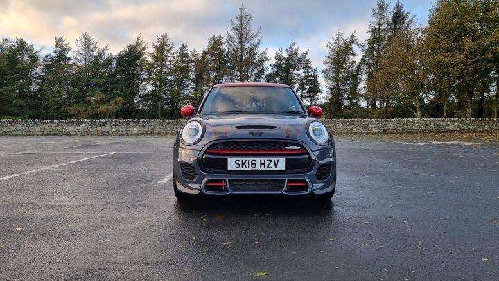 TRD's Mini John Cooper Works F56 - Page 1 - Readers' Cars - PistonHeads UK - The image is a photograph showcasing a black Mini Cooper car with visible modifications, parked in a parking lot. The vehicle features the manufacturer's emblem on its grille and a custom hood with vents, as well as a rear spoiler. The Mini is positioned on the left side of the frame, angled slightly towards the viewer. There are no people or other moving objects in the image, suggesting it was captured during a time of relative stillness. The background reveals an overcast sky and sparse vegetation, with the car partially obscured by a tree.