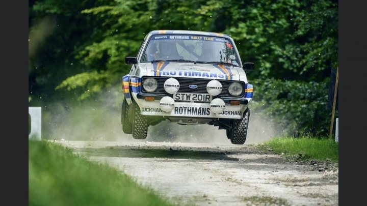 A truck is parked on the side of the road - Pistonheads - The image is a color photograph showcasing a rally car in motion on a dirt road. The vehicle has a distinctive livery with sponsor names and logos, including the text "Purely Motorsport" on its side. It's kicking up dust as it drives, indicating speed. In the background, there are trees and a fence, suggesting a rural or forested area. The driver is visible through the car's windows, focusing intently on the road ahead.