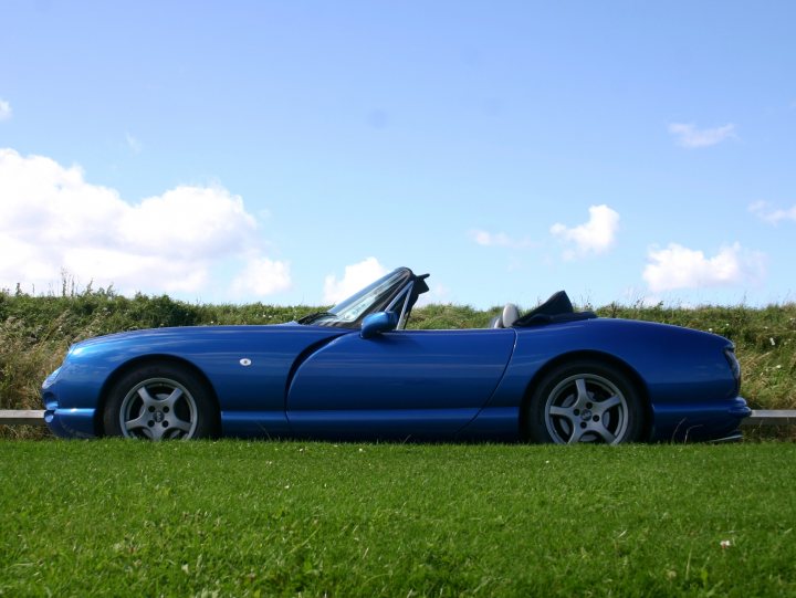 Wow! A sunny day... - Page 1 - North East - PistonHeads - The image depicts a blue sports car parked on a grassy patch. The car is sleek and low-to-the-ground, typical of sports cars, with two doors and a convertible roof. Its design is aggressive with angular lines, and it features alloy wheels. The car appears to be parked during sunrise or sunset, as evidenced by the soft lighting in the sky and the long shadows of the car on the grass. The setting is serene and open, with a slightly undulating lawn leading up to a wooden fence in the background.