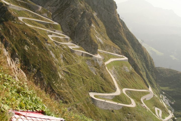 Damage Cobbled Roads Pistonheads - The image showcases a serpentine mountain road that winds its way up a steep, grassy mountain. The road is broad and curvy, appearing to vanish into the distance as it ascends. As one follows the road's twisty path, the view looks down upon the highway and gravel side roads at the bottom of the mountain. The road is surrounded by lush grass and shrubs, offering a verdant green contrast to the gray gravel of the highway. From this high vantage point, one can observe the full extent of the mountain road, which is a testament to human engineering and infrastructure in this rugged natural setting.