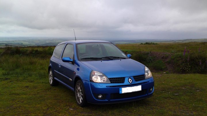 Clio 172 Cup - Page 1 - French Bred - PistonHeads - The image shows a small, blue compact car parked in a field. The vehicle appears to be a hatchback model, positioned at an angle that allows for a view of its front and side. The background reveals a verdant, hilly landscape under an overcast sky. The grassy area around the car is spotted with a few patches of dirt. The image seems to capture a serene setting, possibly in a rural or suburban environment.