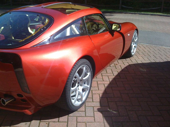 Pistonheads - The image features a red sports car parked on a brick driveway with a manmade fence in the background. The car is an open-topped model with a sleek design, featuring a large Plexiglas skylight in the rear window area. The vehicle has black tires and silver rims, enhancing its sporty appearance. The lighting casts shadows, suggesting that the photograph may have been taken on an overcast day. The overall scene communicates a sense of luxury and ambition.