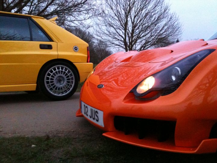 Pistonheads Seconds Endulge - In the image, two sports cars are parked side by side on a street. The car on the left is a vibrant yellow, with a distinctive sloped front and a black soft top roof. It has black rims and a black soft top roof, giving it a sleek appearance. The car on the right is a striking shade of red, with a large front bumper and a unique design with a vent behind the headlight. Both cars are positioned on a street, adjacent to some trees. The bright headlights of the cars suggest they are either parked for the night or prepared for a drive.