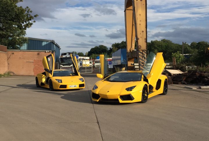 V12 Lambo Photo Thread - Page 3 - Diablo/Murcielago/Aventador - PistonHeads - The image is a photograph showing two yellow sports cars parked in what appears to be an industrial or logistics area. The cars are sleek and low to the ground, with a design that suggests high performance. They are similar in shape but different in size, with the front car appearing larger and the one behind it appearing slightly smaller. The surroundings include concrete pavement, some structures behind the vehicles, and an overcast sky overhead.