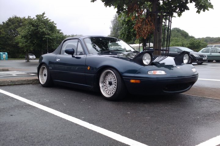 SPOTTED - Lowrider MX5 - Page 1 - South Wales - PistonHeads - The image shows a dark-colored two-door coupe parked in a lot. The vehicle is sleek and appears to be from the 1990s, featuring alloy wheels, tinted windows, and bright headlights. The car is positioned in front of a tree with brownish leaves, suggesting that the photo might have been taken during the autumn season. In the background, there are other cars parked at various angles, giving the scene a busy yet orderly appearance. The overall atmosphere suggests a typical day at a public parking area.