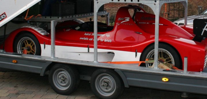 Tiedowns Pistonheads - The image shows a vibrant red and white vintage race car, possibly a single-seater, parked on a trailer. The car's sleek design and visible engine are indicative of its racing heritage. The trailer supporting the car is equipped with a ramp, ready to load or unload the vehicle. In the background, there's a brick wall and another car, suggesting the scene might be taking place at a car show or similar event. The overall atmosphere is one of nostalgia for classic racing cars.