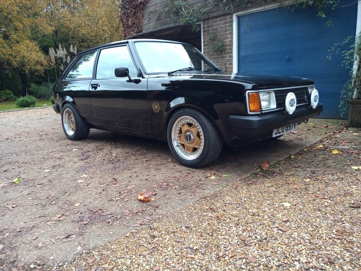 Talbot lotus sunbeam - Page 4 - Classic Cars and Yesterday's Heroes - PistonHeads UK - The image shows a black, two-door hatchback parked on the side of a road. It features a distinctive yellow stripe along its side and unique custom wheels. The car is positioned on what appears to be a driveway or a similar paved area. There are no visible texts or distinctive brands that indicate the make or model of the vehicle. The background includes elements of nature, such as leaves on the ground and trees, suggesting that the photo was taken during autumn or winter.