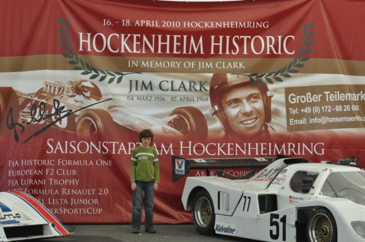 Hockenheimring Clark Pistonheads Jim - The image depicts a boy standing next to a white race car, positioned in front of a stationary banner. The banner is red and features text and artwork related to the Hockenheim Historic, a racing event in memory of Jim Clark. On the banner, there is an illustration of Jim Clark, a renowned Formula 1 driver, in a red race car. The date of the event, April 18, 2010, is indicated along with other details such as tells, racing times, and a section dedicated to FIA Historic Formula 1 cars. The boy appears to be dressed casually, contrasting with the professional setting suggested by the race car and the banner.