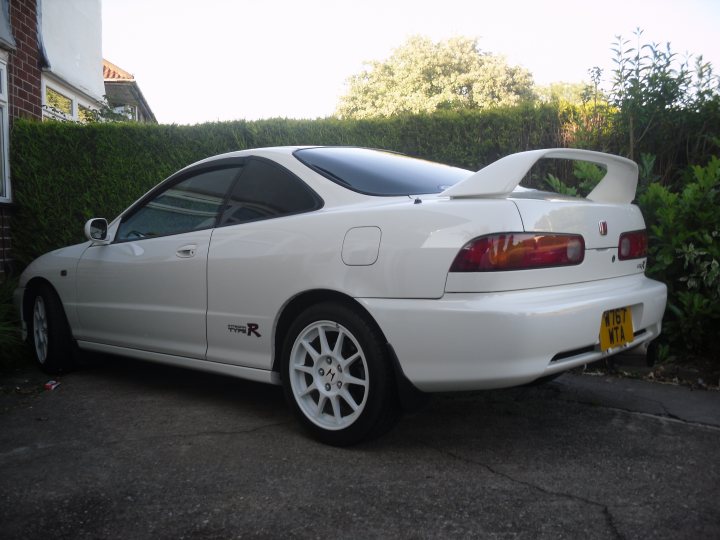 Pistonheads - The image shows a white sports car parked outside a building, facing towards its side. The car is equipped with a rear spoiler and a noticeable angle kit, which gives it a sportier stance. The license plate is clearly visible and has the character string "W70 KTA". The backdrop features a green hedge, and other typical residential elements such as brickwork can be seen. The car's design suggests it's likely a compact or subcompact vehicle, prized for its agility and speed in such a category.