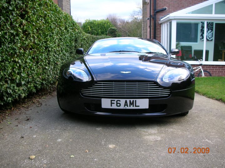 number plates - Page 1 - Aston Martin - PistonHeads - The image features a black Aston Martin F6 AML sports car, viewed from a front three-quarter angle, parked on a paved driveway. The car is in front of a brick house, with green hedges to its left and a white garage door in the background. A bicycle to the right of the car suggests the home's use of alternative transportation methods. The photo also contains a timestamp indicating it was taken on July 2, 2009.