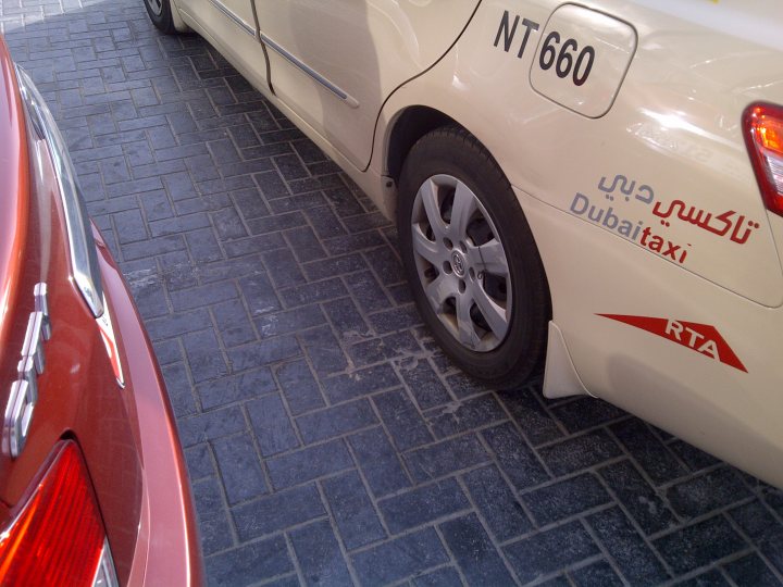 Pistonheads - The image depicts a scene with two cars parked on a brick pavement. In the foreground, there are two vehicles that are the primary focus of the image. The car closest to the camera is a white car with text on it that reads "NT660" and "Dubai TAXI". The other car, partially visible to the left, seems to be red. The brick pavement on which the cars are parked show signs of wear and tear, indicating that this area may be frequented by many vehicles and pedestrians. The overall setting appears to be an outdoor parking lot or a street with limited car traffic.