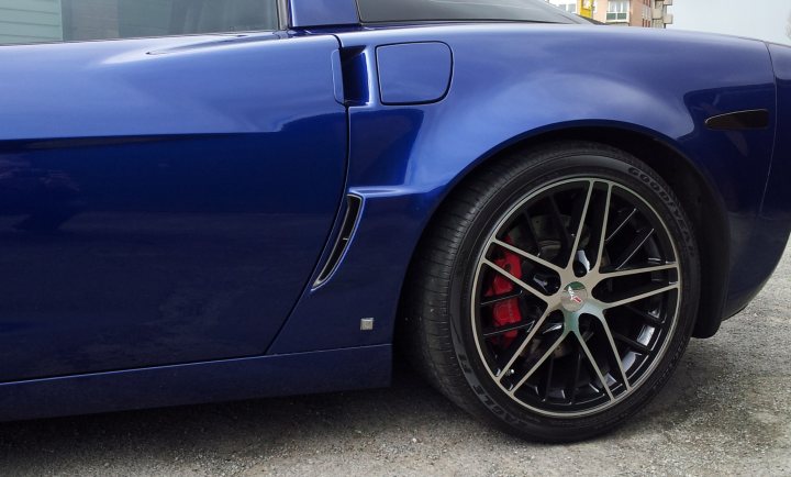 New Wheels - Page 1 - Corvettes - PistonHeads - The image captures a close-up view of a single, stunning sports car. The car's exterior is a deep, glossy blue, reflecting its vibrancy. Its most striking feature is a massive wheel, an integral part of its body, enhancing its sporty appearance. The wheel rims dual colors with orange and black details that provide a nice contrast to the blue of the car.

The car is parked on a street with a garage in the background, suggesting a residential or suburban setting. The focus and perspective of the image emphasize the car's design and details, making it the center of attention. The cityscape lends a sense of scale and urban context to the overall scene.