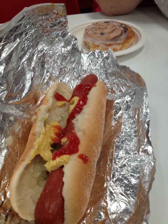 Dirty takeaway pictures Vol 2 - Page 327 - Food, Drink & Restaurants - PistonHeads - The image presents a tantalizing culinary scene. In the center of the frame, a foot-long hot dog is nestled in a bun, generously topped with ketchup and mustard. This hot dog is resting on a piece of foil, which lies on a white cloth-covered dining table. The hot dog's vibrant colors contrast beautifully against the stark white surface of the table. In the background, another dish is visible, featuring a delicious looking dessert or pastry, adding a sweet counterbalance to the savory hot dog. The overall setting suggests a casual dining experience, inviting the viewer into the world of food.