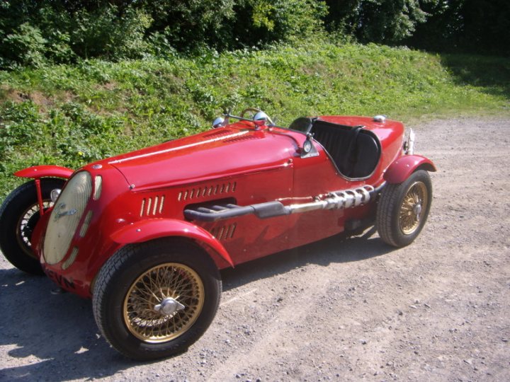 Lets see a picture of your classic(s) - Page 192 - Classic Cars and Yesterday's Heroes - PistonHeads - The image showcases a striking antique model T Ford car parked on a gravel surface. The car's glossy red finish stands out against the natural backdrop composed of a grassy embankment and a dense carpet of trees. Its vintage appearance is accentuated by the visible chrome parts, specifically the bumper and decorative elements, which shine under the light. The old car, despite some signs of wear, retains a sense of charm and nostalgia, and the way it's parked suggests it's ready for a drive.