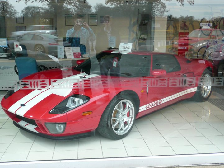 Ford Sale Heath Monks Pistonheads - The image shows a bright red sports car parked inside a showroom with a tiled floor. The car is a Portofino, as indicated by the tag on its windscreen. It has a distinct black stripe running down its side, adding to its sporty appearance. The reflection of a man taking a picture of the car is visible in the showroom's window. The showroom itself has a modern interior, with other vehicles and reflective surfaces creating a spacious and inviting atmosphere.