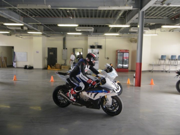 Pistonheads - The image depicts an indoor motorcycle riding facility. It shows a rider in gear atop a motorcycle, practicing among orange traffic cones. Another motorcycle is parked in the background, and a few people are present in the facility. The interior walls are bare, and it appears to be a specialized space for motorcycle training or practice sessions. The lighting in the image gives a clear, well-lit appearance to the room and the motorcycles.