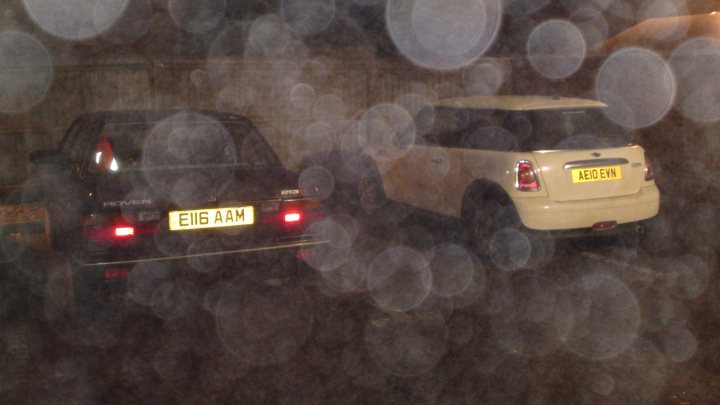 Pistonheads - The image captures a nighttime urban scene featuring two automobiles. In the foreground, there's a black vehicle with the license plate "E16 AAM." The vehicle is stationary, and the brake light is red, indicating it has come to a stop. In the background, there's a white hatchback with the license plate "A18 EV0." The hatchback appears to be parked in a lot or space adjacent to the black vehicle. The surroundings are blurred, adding emphasis to the vehicles. Public lighting can be seen, illuminating the area in a soft glow. The scenario suggests a moment of inactivity or stillness in an everyday cityscape.