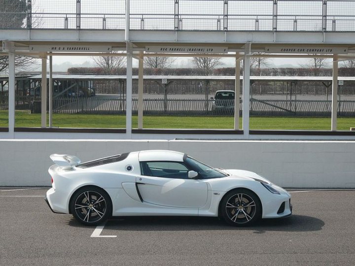 At Goodwood today..... - Page 1 - Goodwood Events - PistonHeads - The image features a white two-door sports car, likely a convertible given its visibly open roof, parked in a designated parking bay. The vehicle is positioned next to a protective barrier, possibly part of a trackside viewing area. The setting suggests a motorsport facility or a track, possibly in a location where English is spoken due to the English road sign visible in the background. The background scene includes empty stands and a truck, hinting at the track being unused at the moment of the photograph.