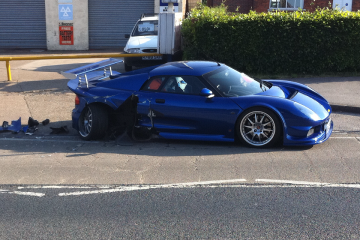 Lets see your Noble's - Page 1 - Noble - PistonHeads - This image captures a scene of accidental damage. A dark blue sports car is prominently displayed on the road, with its front end crumpled and parts of the bodywork scratched and dented. The car is positioned on some sort of median between lanes of traffic, with vehicles on both sides visible in the background. The setting appears to be an outdoor urban area, with taller structures in the distance and shrubbery lining the roadside. There are no people in sight, and the focus of the image is solely on the condition of the car.