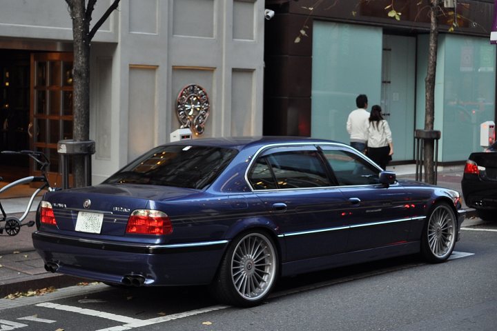 Rarities Spotted Supercars Pistonheads - The image shows a street scene featuring a parked dark blue luxury car at the side of the street. In the background, there is a building with a unique architectural feature, a tree extending from the sidewalk, and pedestrians walking on the sidewalk. The car is parked adjacent to a parking meter, and in the near background, there are other cars. The lighting in the image appears to be natural, suggesting it might be daytime.