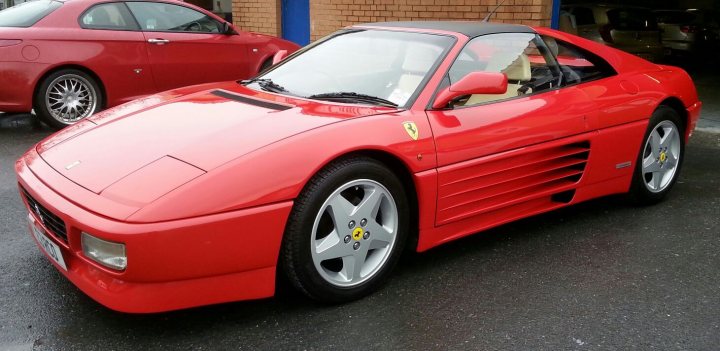 Ferrari 348 chit chat - Page 1 - Ferrari V8 - PistonHeads - The image features a bright red Ferrari on a wet asphalt surface. The car is parked and has a classic design with large wheel arches and a distinctive nose. On the side of the car is the Ferrari emblem and it also has the traditional yellow callipers. There are two other cars in the background, suggesting a parking area or a showroom setting.