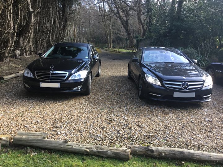 CL vs S class?  - Page 2 - Mercedes - PistonHeads - The image shows two sleek black Mercedes-Benz cars parked side by side on a gravel driveway. The cars are modern, with a distinctive Mercedes-Benz grille design. Behind the vehicles, a line of trees and shrubs suggest a residential or park setting. The sky is overcast, providing soft, diffused lighting. The image appears to be taken during the day. The scene is neat and orderly, with the cars parked correctly and no visible clutter or debris.