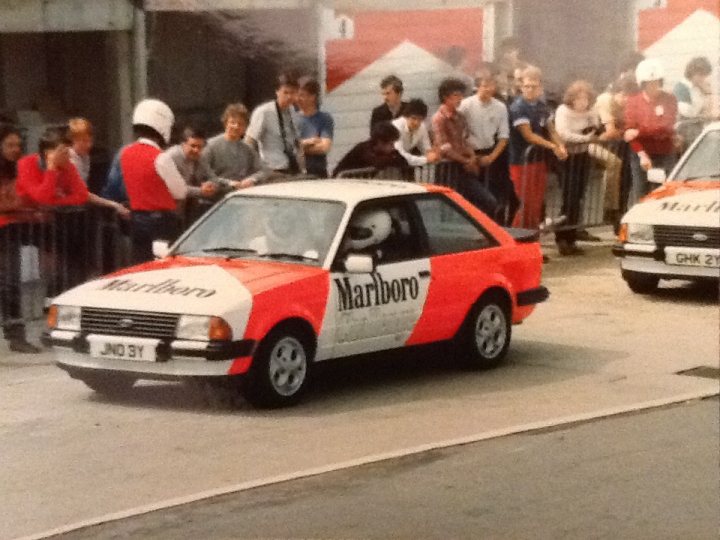 A group of people standing in front of a truck - Pistonheads - In the center of the image, a red and white Marlboro race car is parked, its hood up, possibly undergoing maintenance or waiting in the pit lane. Surrounding the racing car are several enthusiasts, their interest piqued by the spectacle. To the right, there's another Marlboro-liveried race car, parked in a similar manner. The setting appears to be a racing circuit, with concrete barriers and a fence in the background, hinting at the excitement that presumably follows these cars when they are on the track.