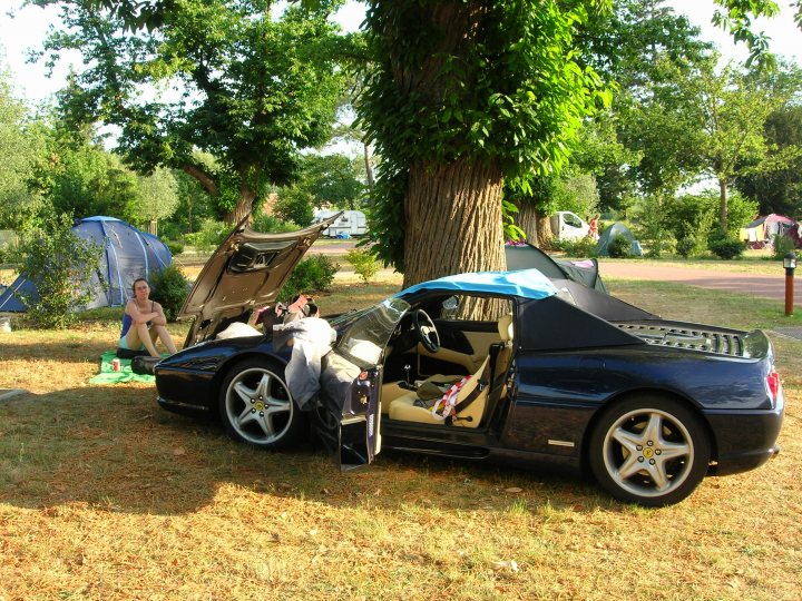 Bruges Park Pistonheads - In the image, a black convertible sports car is parked on a grassy area with a dirt path nearby. The car's door is open, revealing two roadside assist kits inside. The car appears to be the main subject of the image. In the background, there is a tent and tents of different sizes, suggesting a campsite or outdoor event. Additionally, there's a person sitting on the grass next to the car, taking a break from the outing. A tree is also visible in the scene.