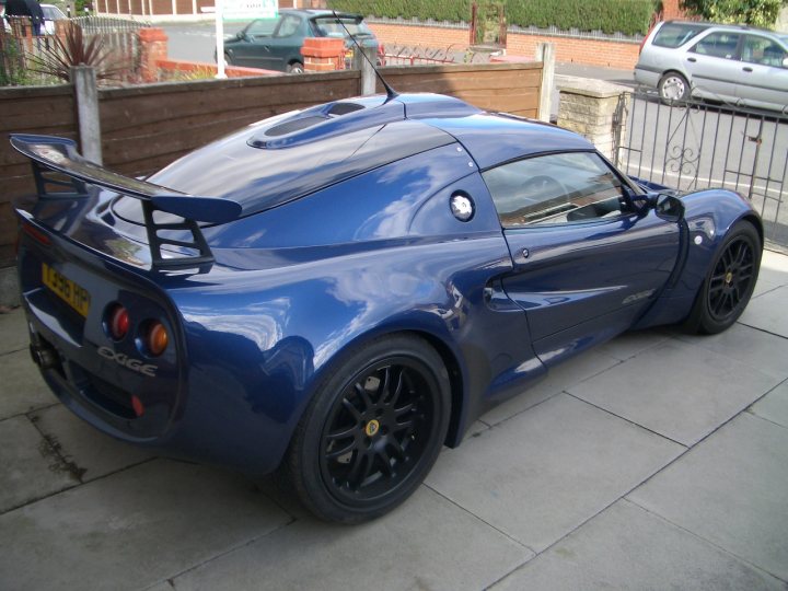 Trying to locate my old Exige... - Page 1 - Elise/Exige/Europa/340R - PistonHeads - The image showcases a blue sports car, parked near a fence and driveway. The car is sleek and shiny, featuring a prominent rear wing and sporty side mirrors. It's parked on a clean, paved driveway, which appears to be wet. The car's color contrasts with the muted tones of the surroundings.